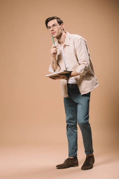 Pensive man in glasses holding notebook and pen on beige background — Stock Photo