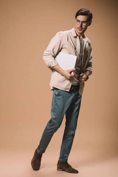 Hombre serio en gafas que sostienen el ordenador portátil sobre fondo beige - foto de stock