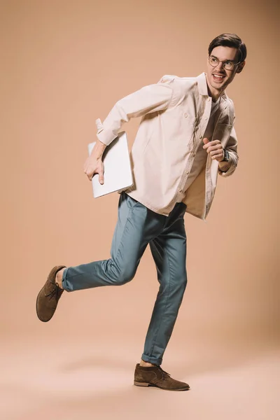 Homem feliz em óculos correndo com laptop no fundo bege — Fotografia de Stock