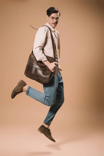 Handsome man in glasses jumping with bag on beige background — Stock Photo