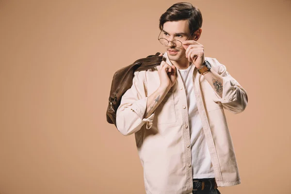 Hombre guapo tocando gafas mientras está de pie con bolsa aislada en beige - foto de stock