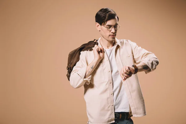 Serious man in glasses looking at watch while standing with bag isolated on beige — Stock Photo