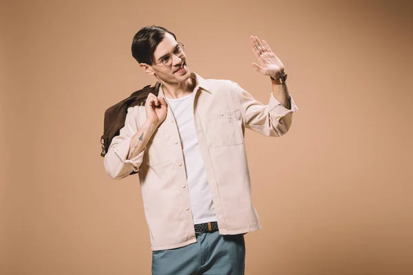 Hombre guapo en gafas agitando la mano mientras sostiene el bolso aislado en beige - foto de stock