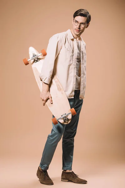 Serious man in glasses holding skateboard while standing on beige background — Stock Photo