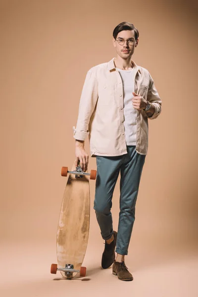 Уверенный в себе человек в очках, держа скейтборд, стоя на бежевом фоне — стоковое фото
