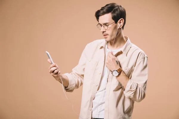 Красивый мужчина в очках, смотрящий на смартфон, слушая музыку в наушниках, изолированных на бежевом — стоковое фото