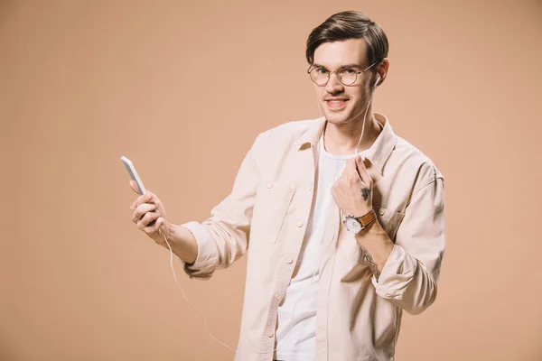 Homem sorridente em óculos segurando smartphone e ouvindo música em fones de ouvido isolados em bege — Fotografia de Stock