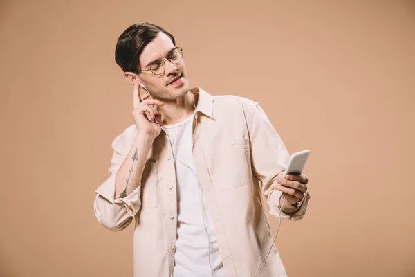 Handsome man in glasses holding smartphone and listening music in earphones with closed eyes isolated on beige — Stock Photo