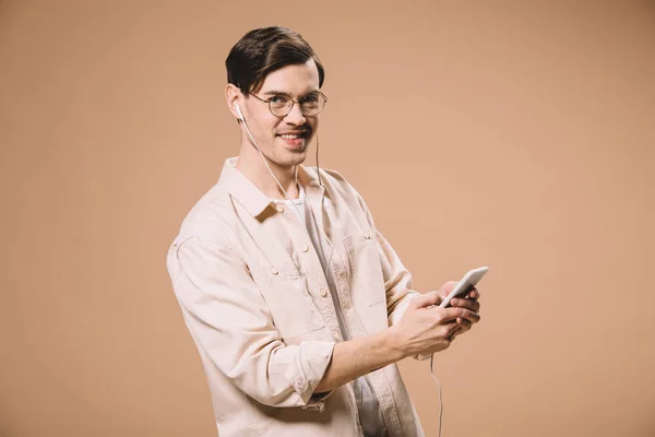 Веселий чоловік в окулярах тримає смартфон і слухає музику в навушниках ізольовано на бежевому — стокове фото