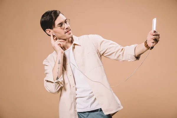 Bel homme en lunettes regardant smartphone tout en écoutant de la musique isolée sur beige — Photo de stock