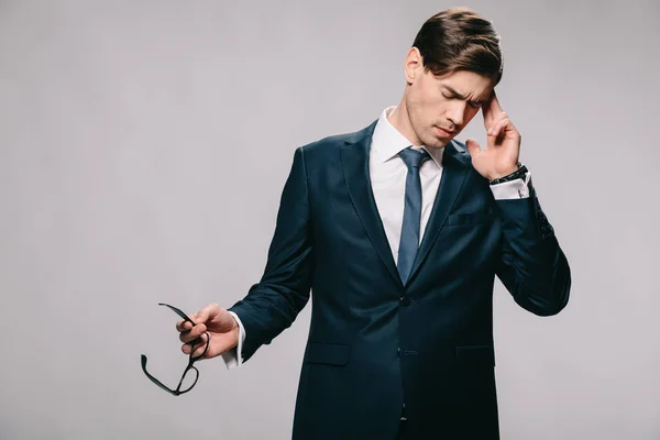 Guapo hombre de negocios con dolor de cabeza y sosteniendo gafas aisladas en gris - foto de stock