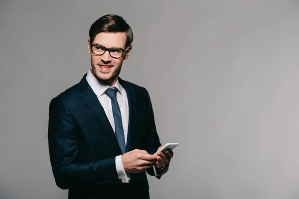 Cheerful businessman using smartphone while standing in suit on grey background — Stock Photo