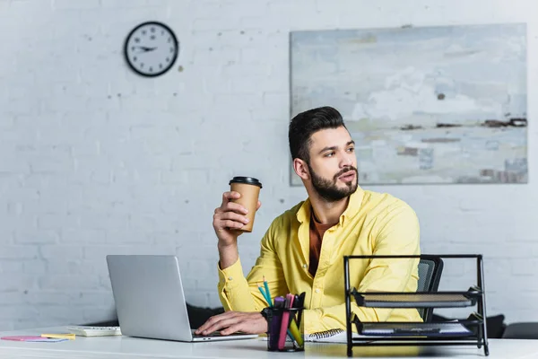 Homme d'affaires barbu tenant tasse jetable et regardant loin sur le lieu de travail — Photo de stock