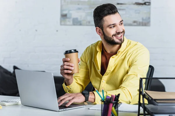 Empresário sorridente segurando copo de papel, usando laptop e olhando para longe — Fotografia de Stock