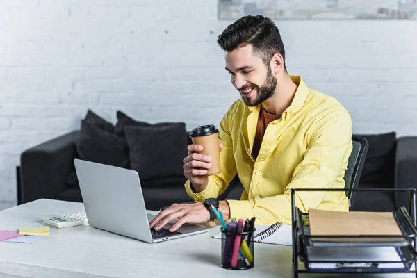 Alegre hombre de negocios sosteniendo la taza de papel y mirando la pantalla del ordenador portátil - foto de stock