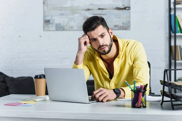 Thoughtful bearded businessman in yellow shirt looking at camera and using laptop — Stock Photo