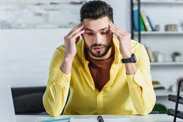 Thoughtful bearded businessman in yellow shirt touching head at workplace — Stock Photo