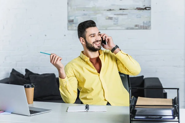 Alegre hombre de negocios barbudo sosteniendo el lápiz y hablando en el teléfono inteligente - foto de stock