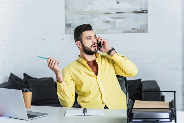 Surprised bearded businessman holding pencil and talking on smartphone — Stock Photo