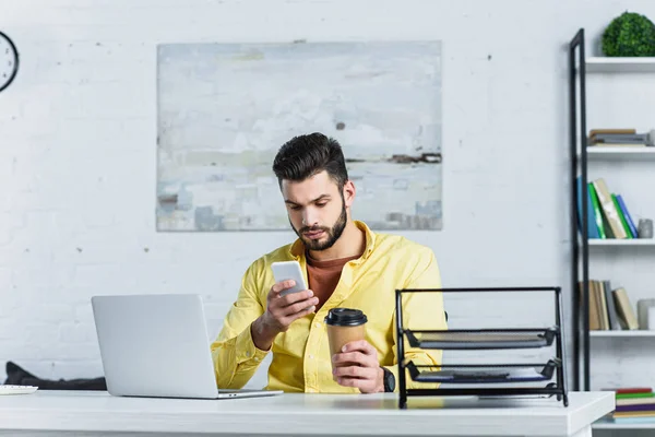 Focused bearded businessman looking at smartphone and holding paper cup — Stock Photo