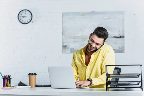 Hombre de negocios barbudo sonriente con camisa amarilla hablando en el teléfono inteligente en el lugar de trabajo - foto de stock