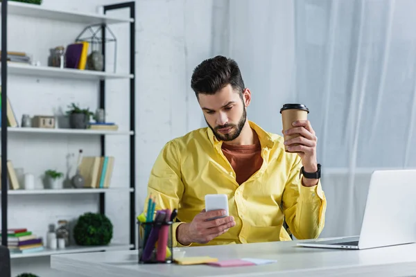 Empresario barbudo enfocado sosteniendo una taza de papel y mirando el teléfono inteligente - foto de stock