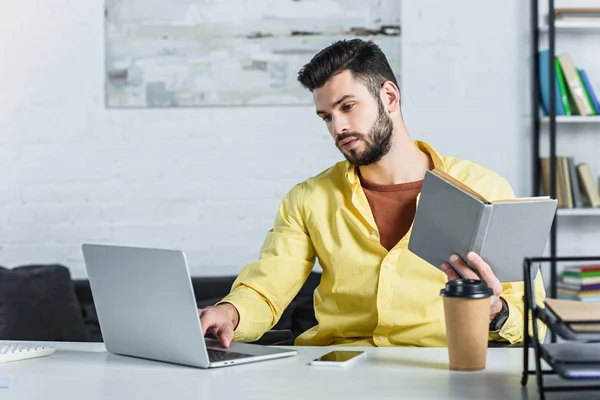 Focused bearded businessman typing and holding book at office — Stock Photo