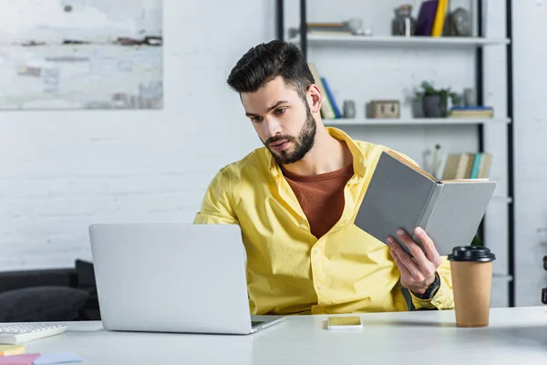 Homem de negócios barbudo concentrado olhando para laptop e livro segurando — Fotografia de Stock