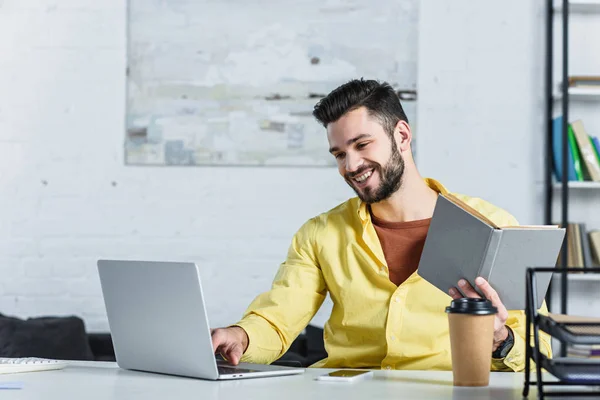 Lächelnder bärtiger Geschäftsmann blickt auf Laptop und hält Buch am Arbeitsplatz — Stockfoto