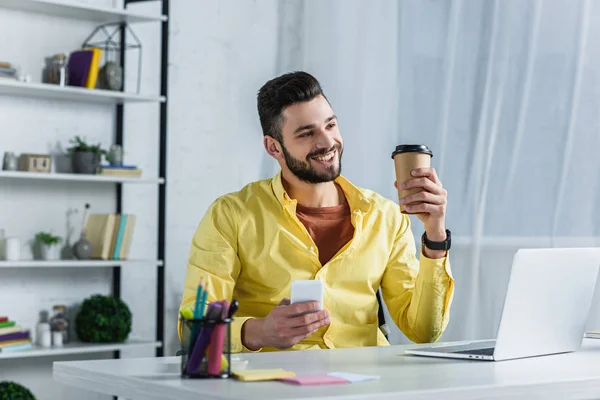 Uomo d'affari sorridente in camicia gialla che tiene tazza di carta e smartphone e distoglie lo sguardo — Foto stock