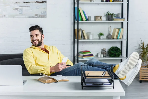 Allegro uomo d'affari barbuto che tiene lo smartphone e distoglie lo sguardo dal posto di lavoro — Foto stock