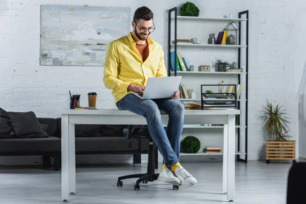 Uomo d'affari barbuto sorridente in occhiali digitando nel computer portatile e seduto sulla scrivania — Foto stock