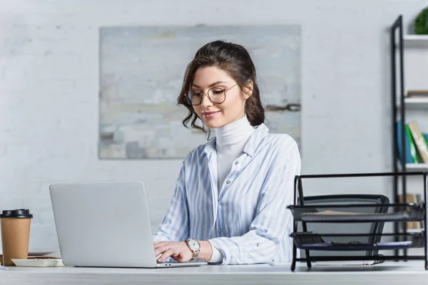 Femme d'affaires souriante en lunettes tapant sur ordinateur portable sur le lieu de travail — Stock Photo