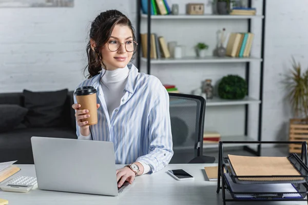 Привлекательная деловая женщина держит бумажный стаканчик и смотрит в сторону на рабочем месте — стоковое фото