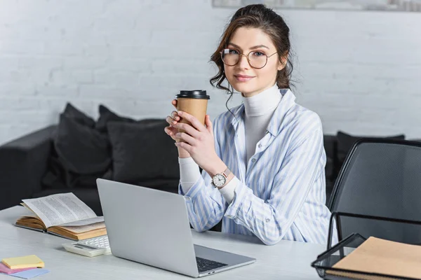 Attractive businesswoman holding paper cup, using laptop and looking at camera — Stock Photo