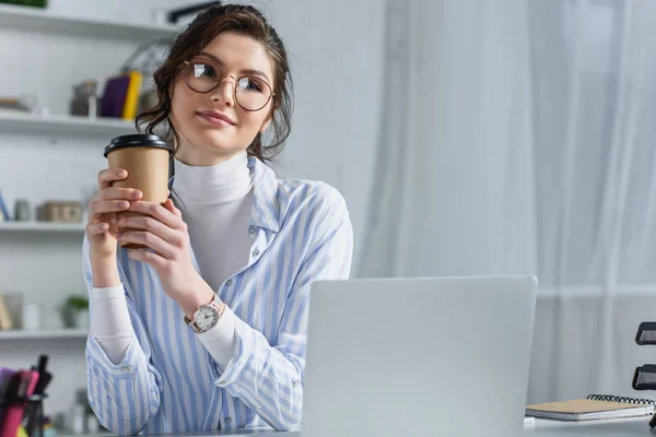 Beautiful businesswoman holding paper cup and looking away at workplace — Stock Photo