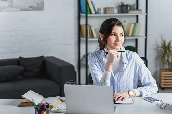 Atractiva mujer de negocios sosteniendo la pluma, utilizando el ordenador portátil y mirando hacia otro lado - foto de stock