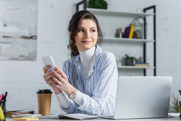 Attractive businesswoman holding smartphone and  looking away at office — Stock Photo