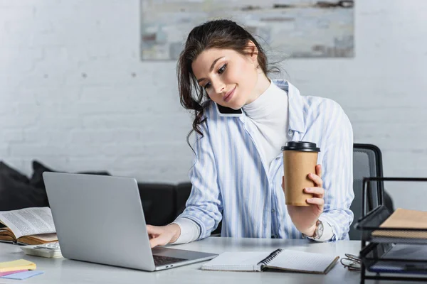 Hermosa mujer de negocios sosteniendo taza de papel y hablando en el teléfono inteligente en la oficina - foto de stock