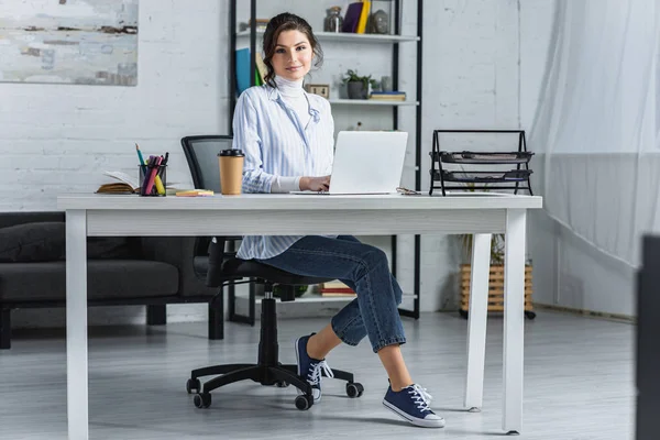 Femme gaie utilisant sur ordinateur portable tout en étant assis dans le bureau moderne — Photo de stock