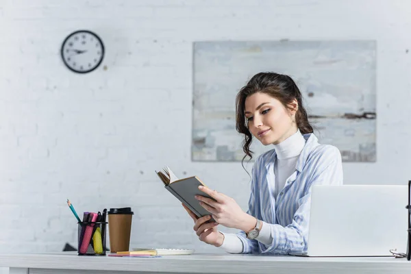 Happy woman rstudying withding book near laptop in modern office — Stock Photo