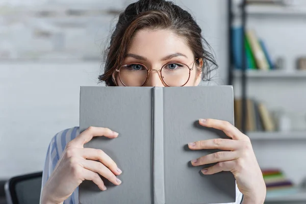 Mulher em óculos cobrindo rosto com livro no escritório moderno — Fotografia de Stock