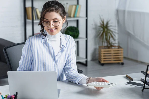 Привлекательная женщина в очках держит калькулятор рядом с ноутбуком в современном офисе — стоковое фото