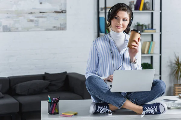 Donna allegra in cuffia con tazza di carta e seduta sul tavolo con computer portatile — Foto stock