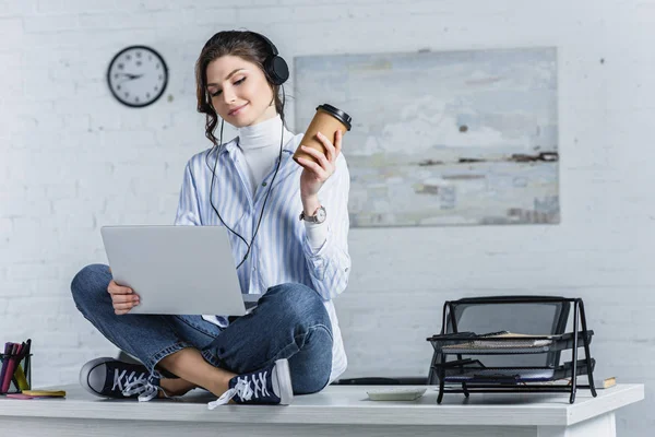 Привлекательная женщина в наушниках держит бумажную чашку и сидит на столе с ноутбуком — стоковое фото
