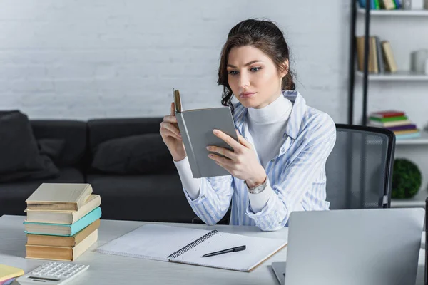 Mujer molesta estudiando con libro cerca de la computadora portátil en la oficina moderna - foto de stock