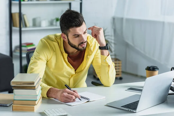 Hombre guapo escribiendo en el cuaderno y mirando el ordenador portátil en la oficina moderna - foto de stock