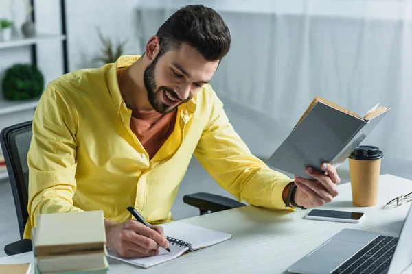 Cheerful man writing in notebook and holding book in hand in modern office — Stock Photo