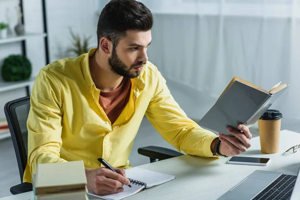 Bell'uomo che studia con libro e computer portatile in ufficio moderno — Foto stock