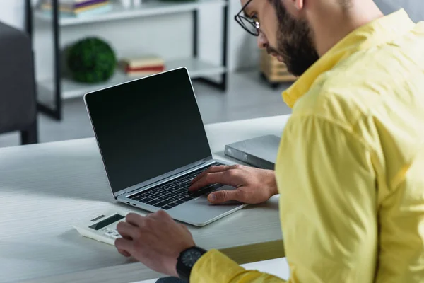 Homem barbudo olhando calculadora perto de laptop com tela em branco no escritório moderno — Fotografia de Stock